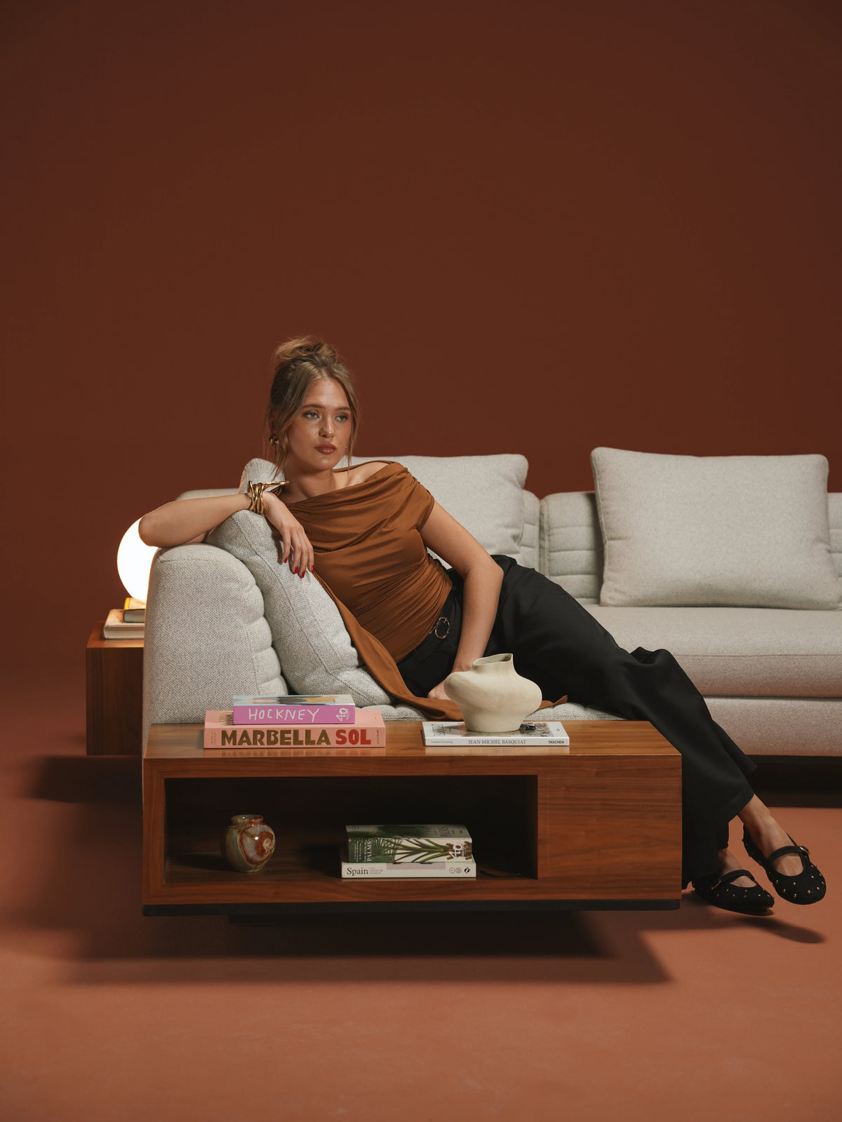 Woman seated on a beige Edge sofa with feather back cushions, inverted seam stitching, and an integrated wooden side table.