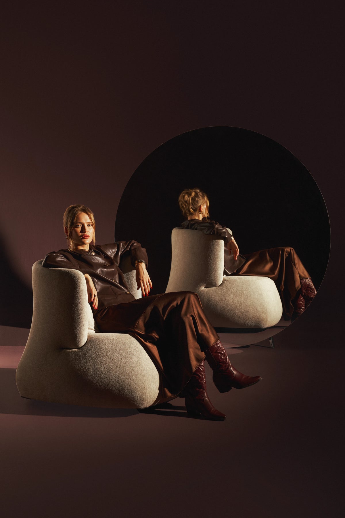 Woman seated in the cream Belly armchair, showing its wraparound backrest and rounded profile beside a circular mirror.