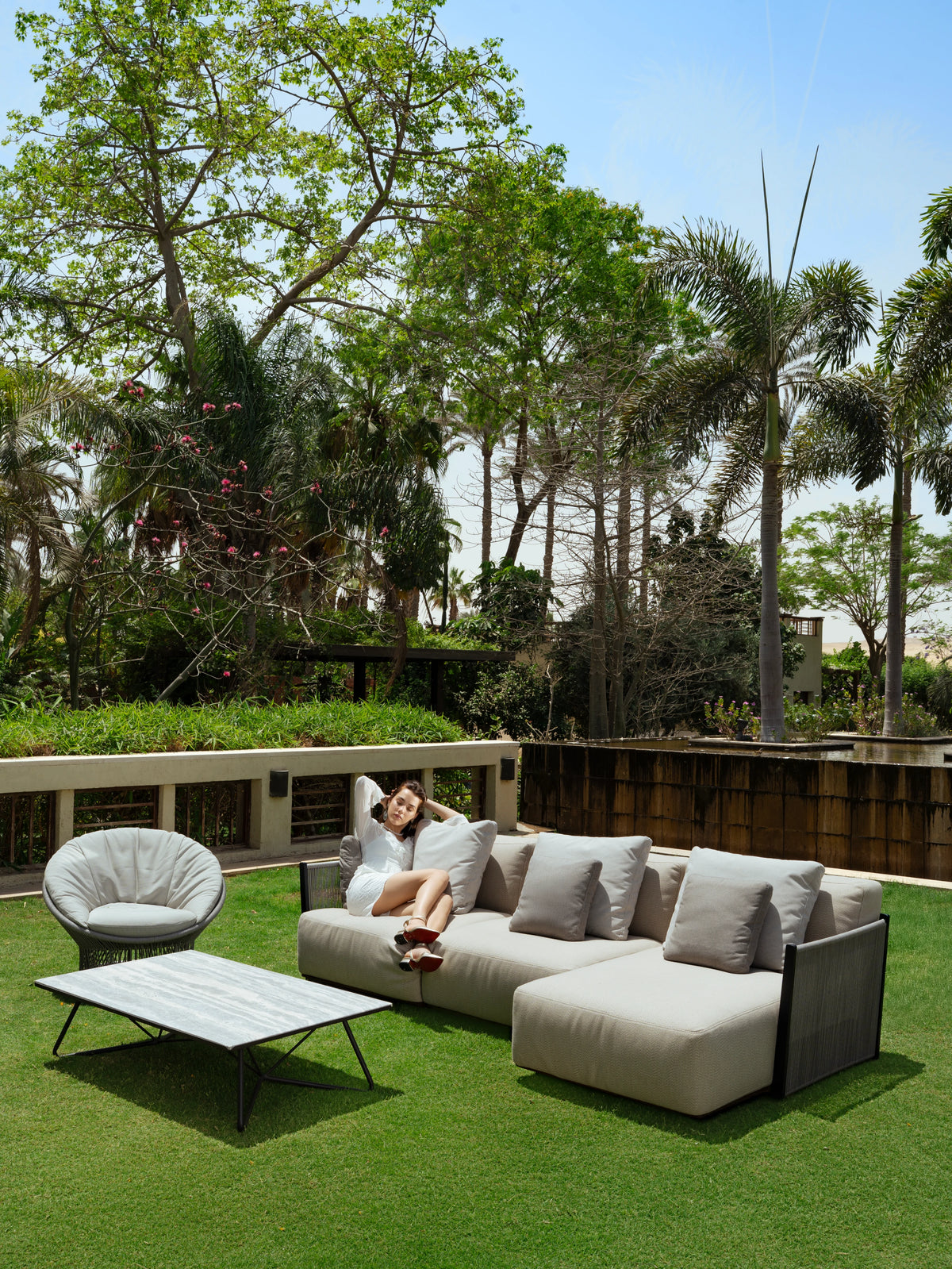 Woman relaxing on a beige Caribbean outdoor sectional sofa, arranged with a Sun armchair and Geo coffee table in a garden.