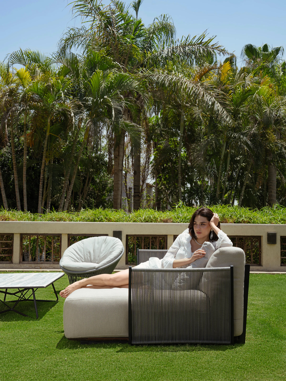 Side view of a woman lounging on the beige Caribbean outdoor sofa, showing its grey vertical rope detailing and chaise section.