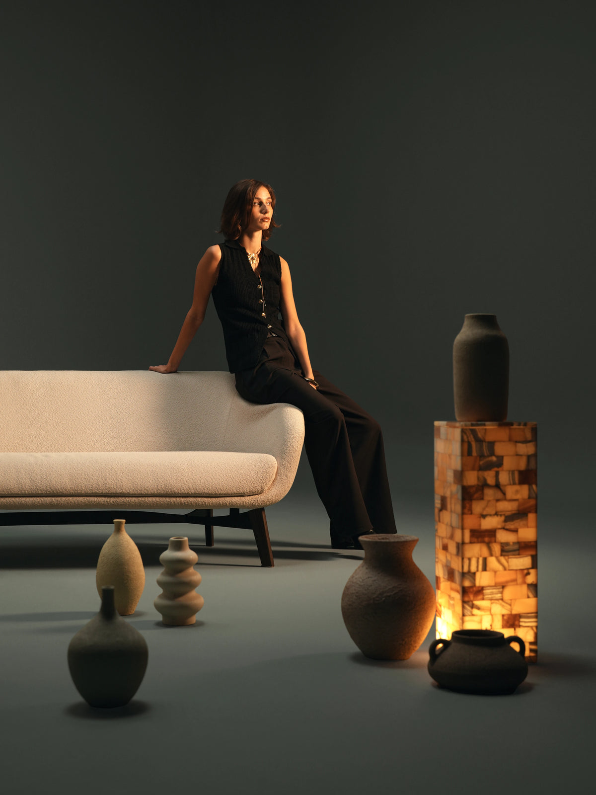 Woman seated on the curved backrest of the compact beige Darby sofa, styled with ceramic vessels in a studio setting.