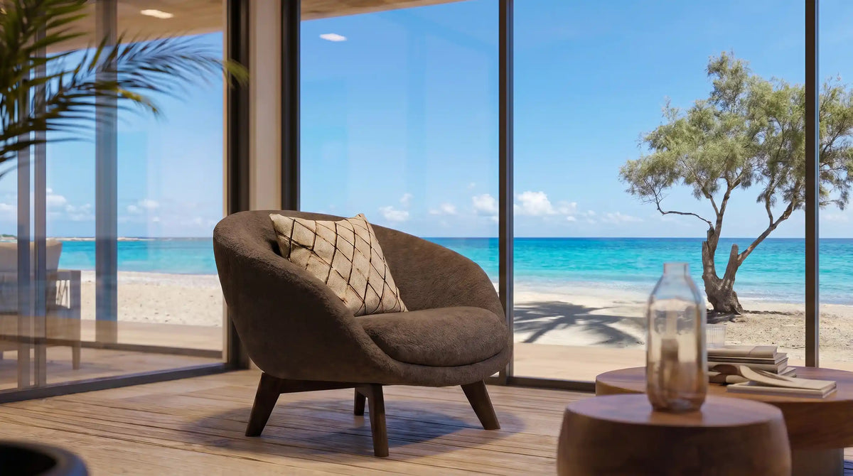 Gray upholstered Darby armchair with a curved backrest, loose cushion, and solid wood legs beside floor-to-ceiling windows overlooking the beach.