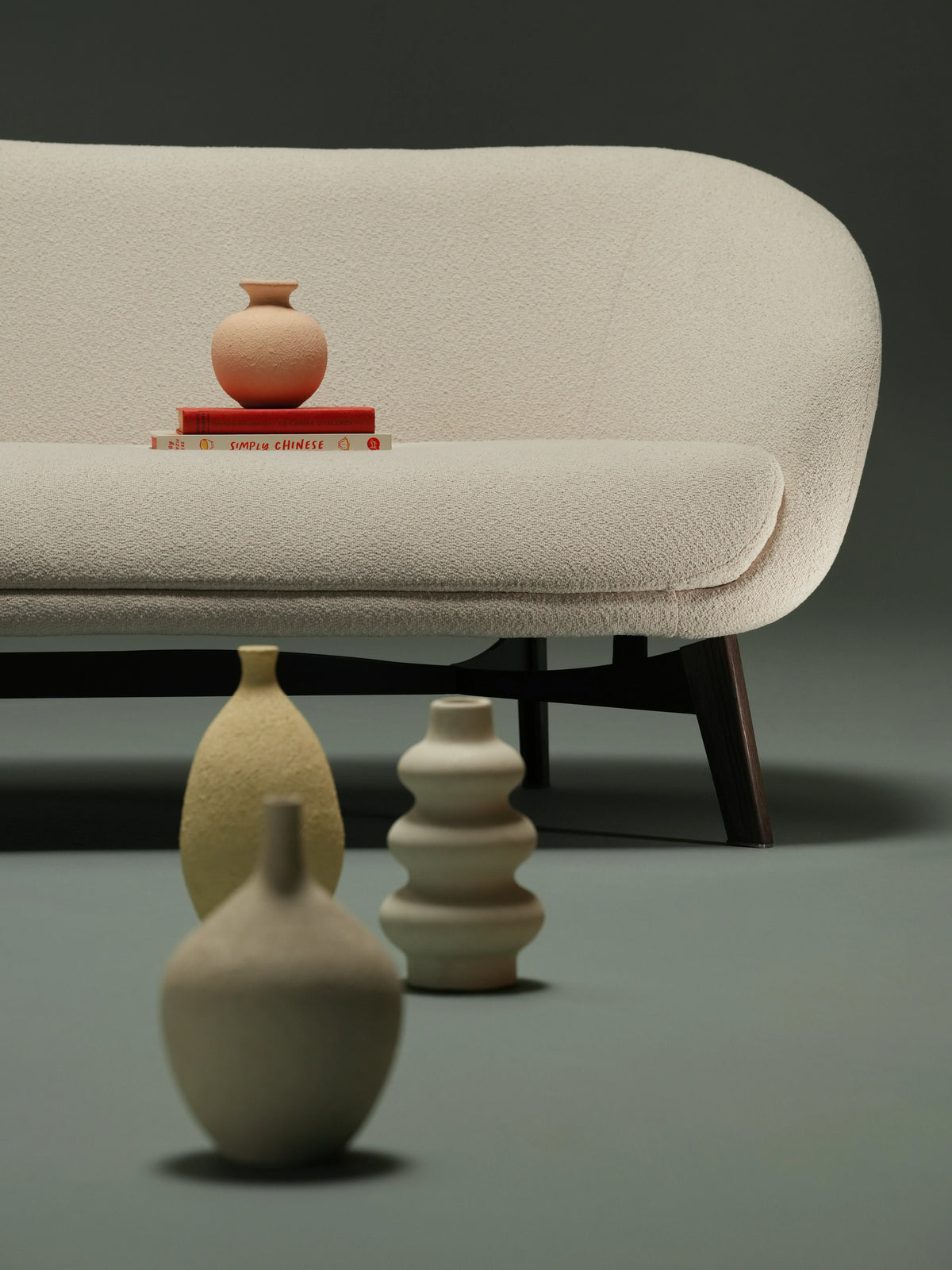 Woman seated on the curved backrest of the compact beige Darby sofa, styled with ceramic vessels in a studio setting.