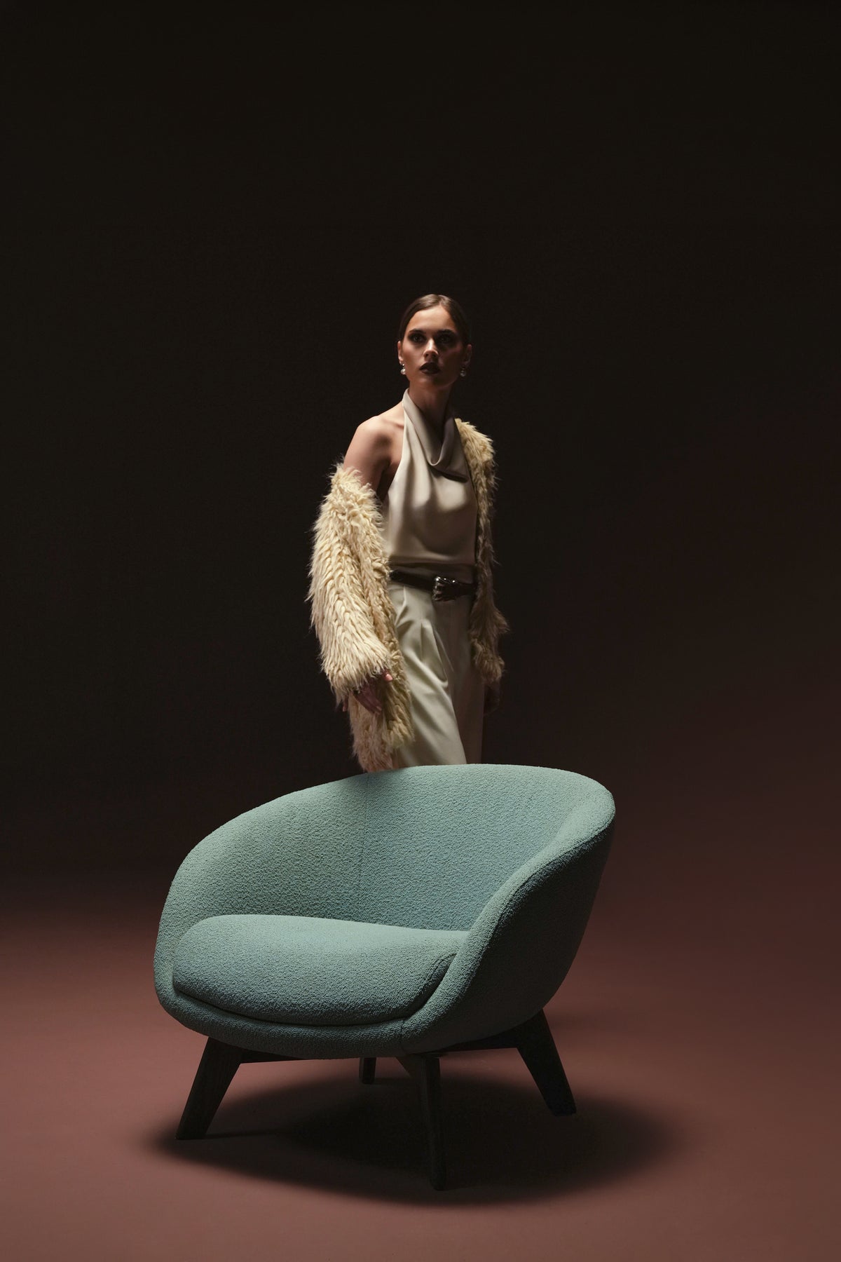 Teal Darby armchair with a curved upholstered back, supportive seat cushion, and dark solid wood spider-leg base, with a woman standing behind it.
