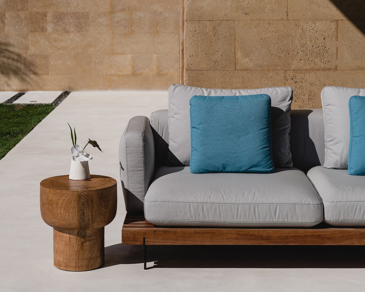 Grey Dock outdoor sofa with blue cushions and a solid teak platform, arranged against a stone wall on a shaded patio.