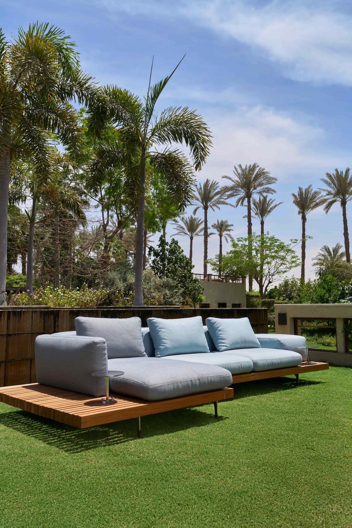 Blue Dock outdoor sofa with broad upholstered armrests and a wide solid teak platform on a garden lawn.