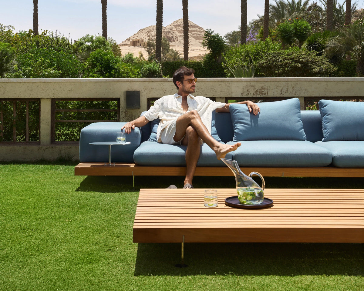 Man seated on a blue Dock outdoor sofa with a wide slatted teak platform and matching Dock table in a garden.