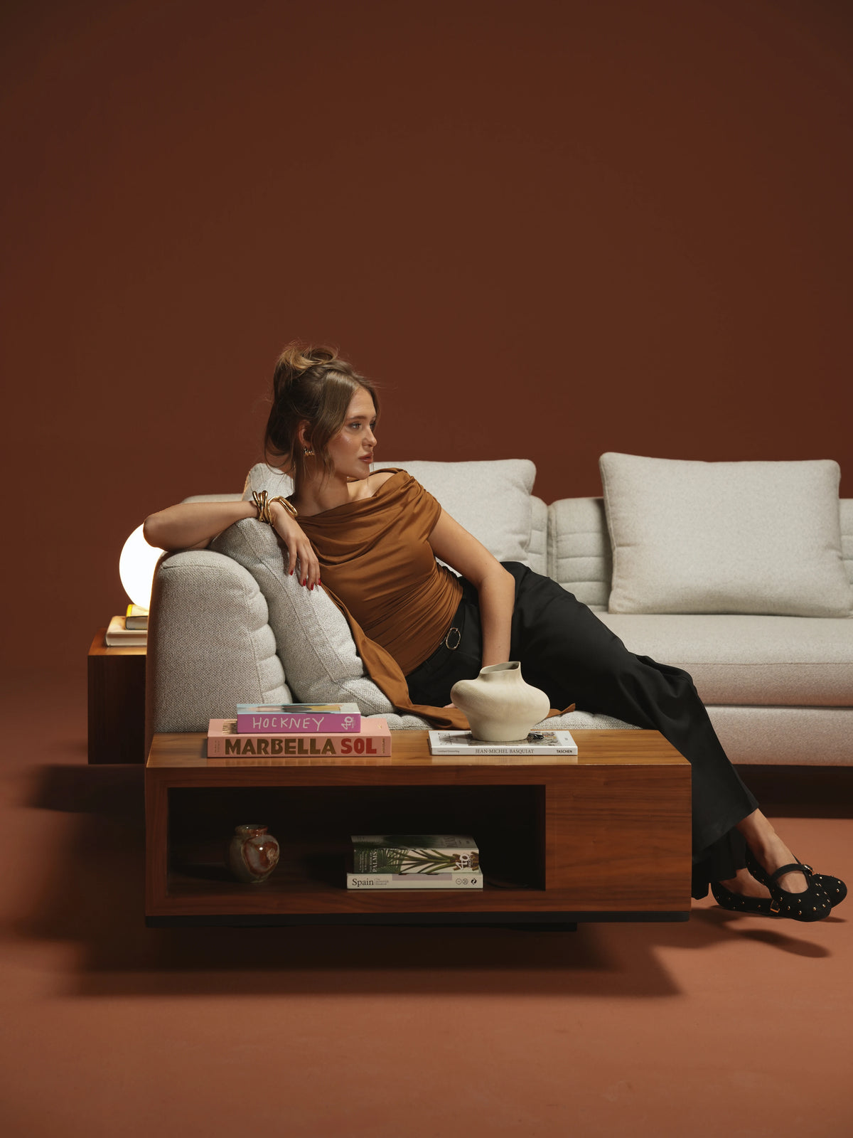 Woman reclining on a beige Edge sofa with inverted seam stitching beside an integrated wooden table with open storage.