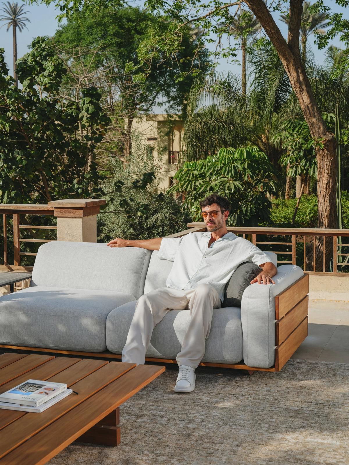 Man seated on the grey Fatty outdoor sofa with bombé-style arms, a solid teak frame, and the matching Fatty table.