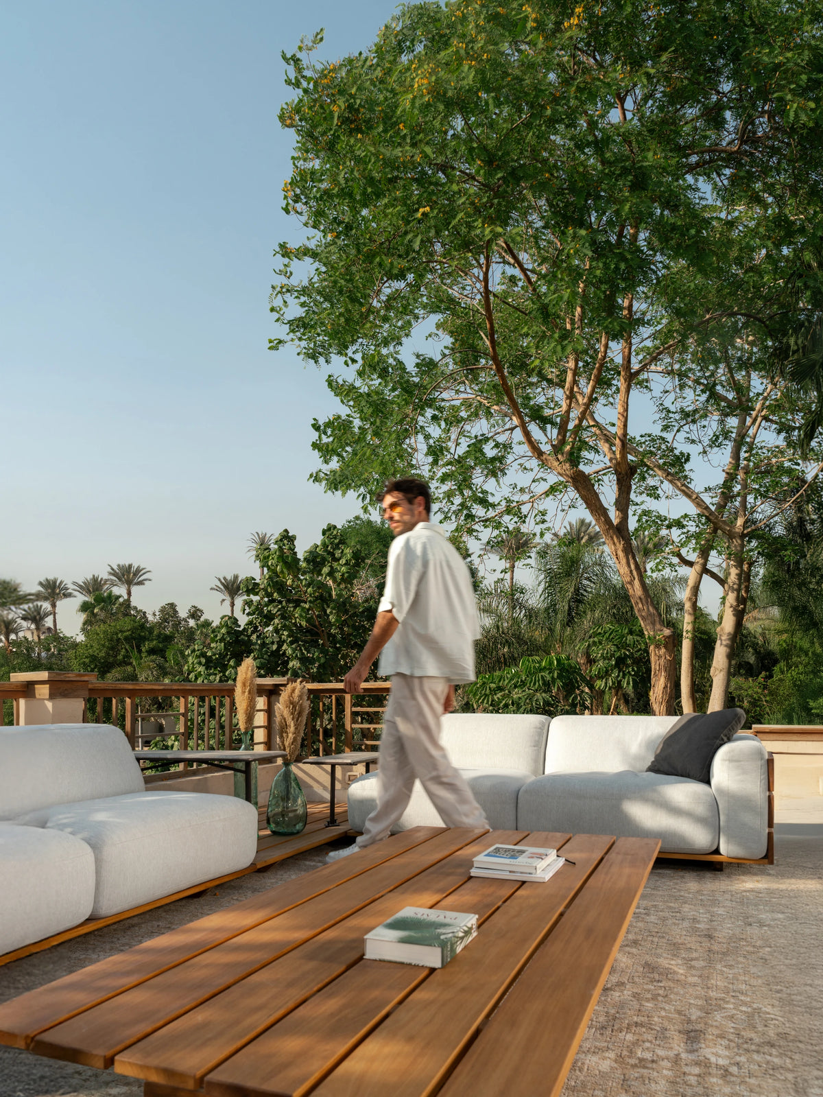 Fatty outdoor sofa with generous grey upholstered cushions and solid teak wood side panels, styled on a terrace garden with Fatty coffee table and model walking by.