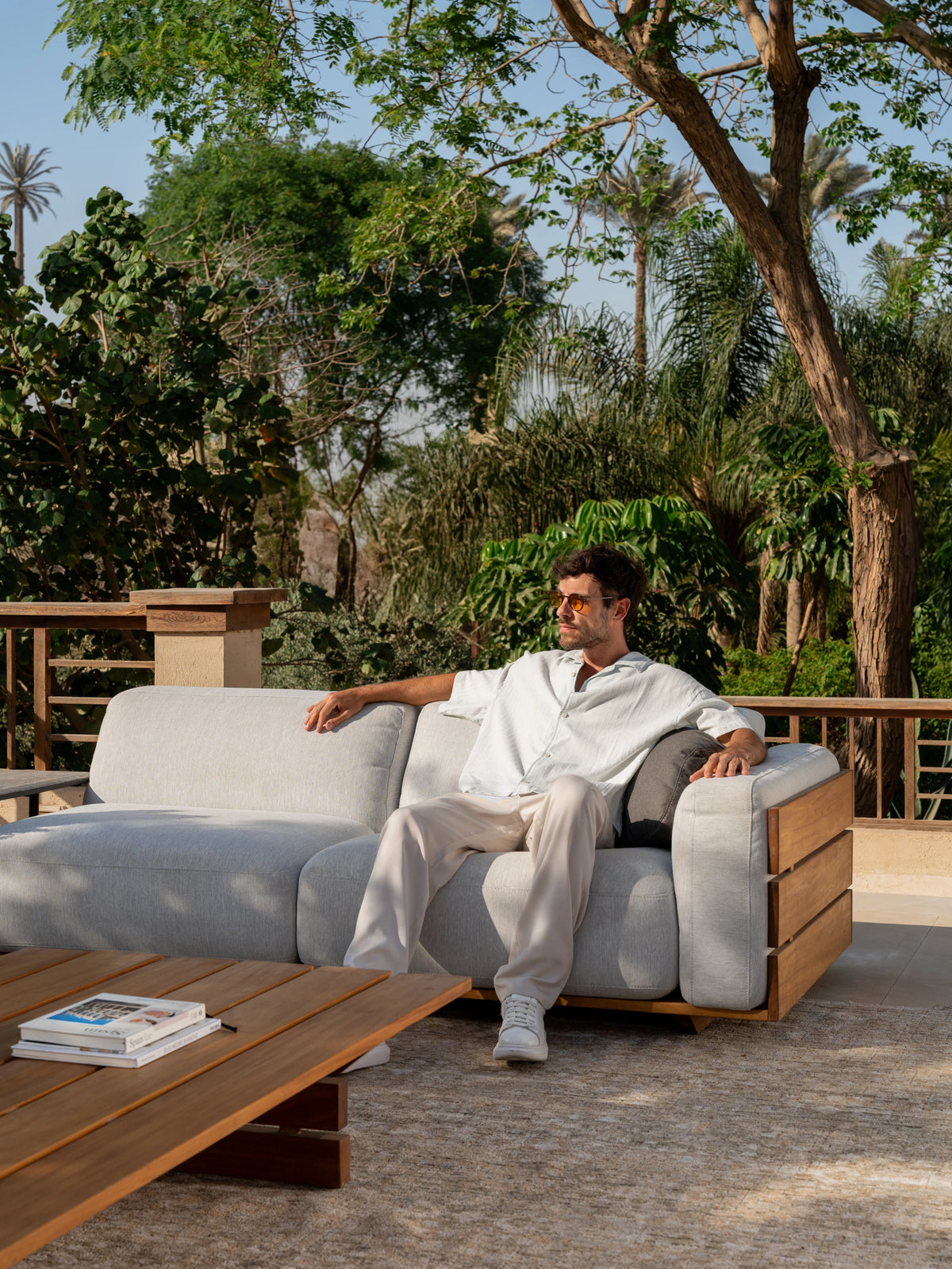 Model seated on Fatty sofa with grey upholstered backrests, showcasing teak wood side framing and deep, grounded proportions in an outdoor setting.