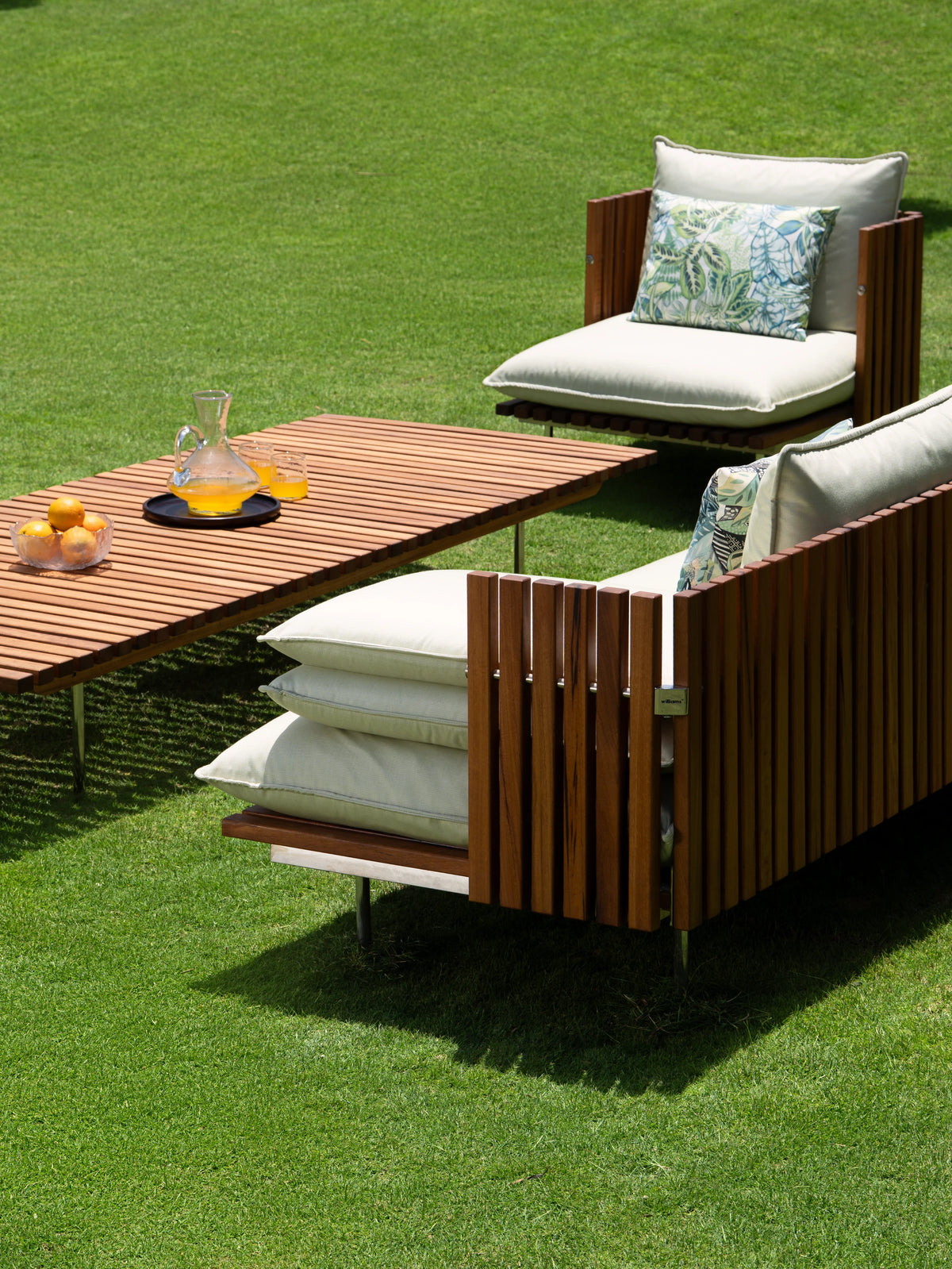 Two Fence outdoor armchairs with vertical teak slat frames and cream cushions, arranged around a matching slatted teak coffee table.