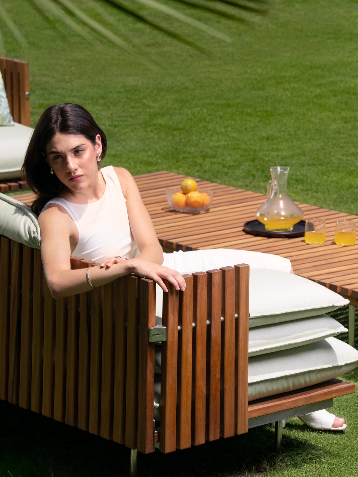 Woman seated in a cream Fence outdoor armchair with vertical teak slats, beside a matching teak coffee table set with drinks and fruit.