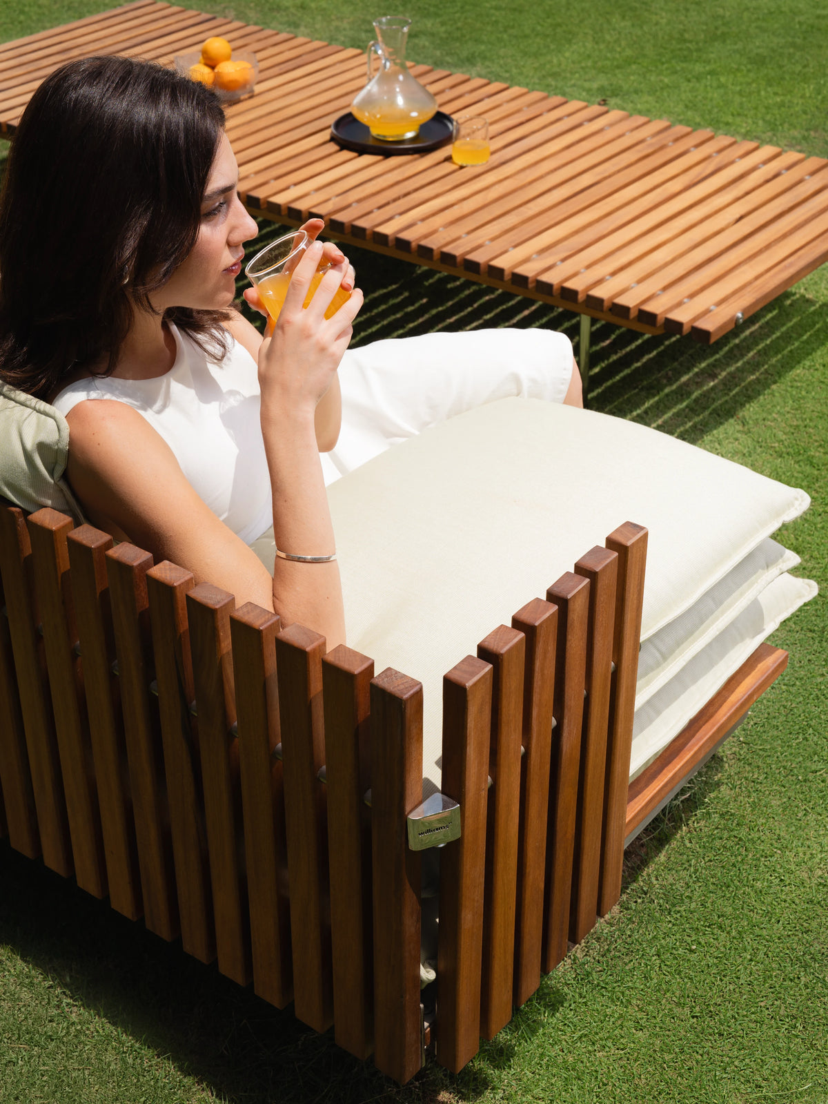 Woman drinking from a glass while seated in a cream Fence outdoor armchair, with the matching slatted teak coffee table behind her.