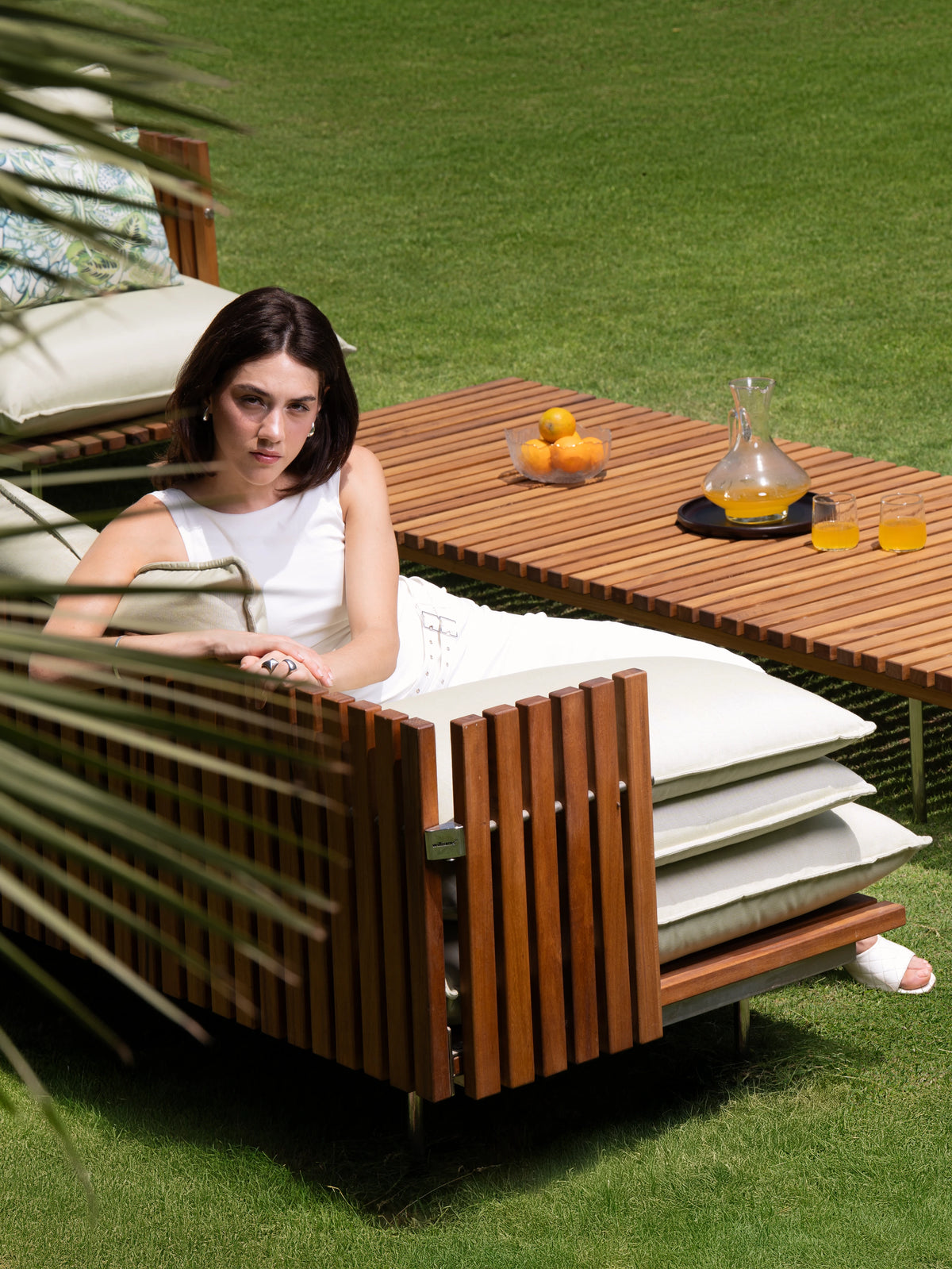 Woman seated in a Fence outdoor armchair with cream cushions and a vertical teak slat frame, beside the matching coffee table.
