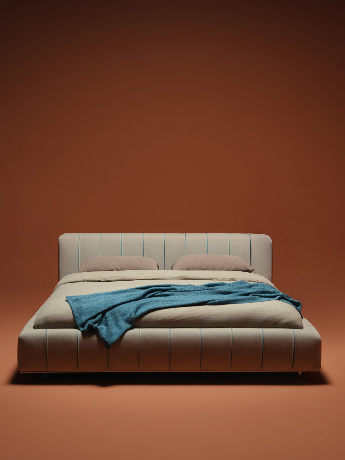 Front view of the Lush bed with a recessed mattress, rounded beige upholstered frame, blue seam detailing and a blue throw.