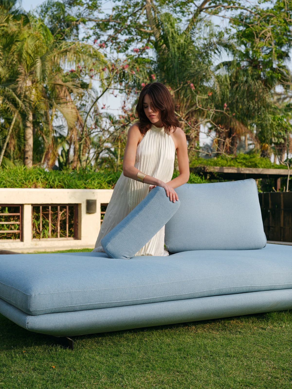Woman repositioning a back cushion on the light blue Island outdoor sofa, demonstrating its flexible seating arrangement in a garden.