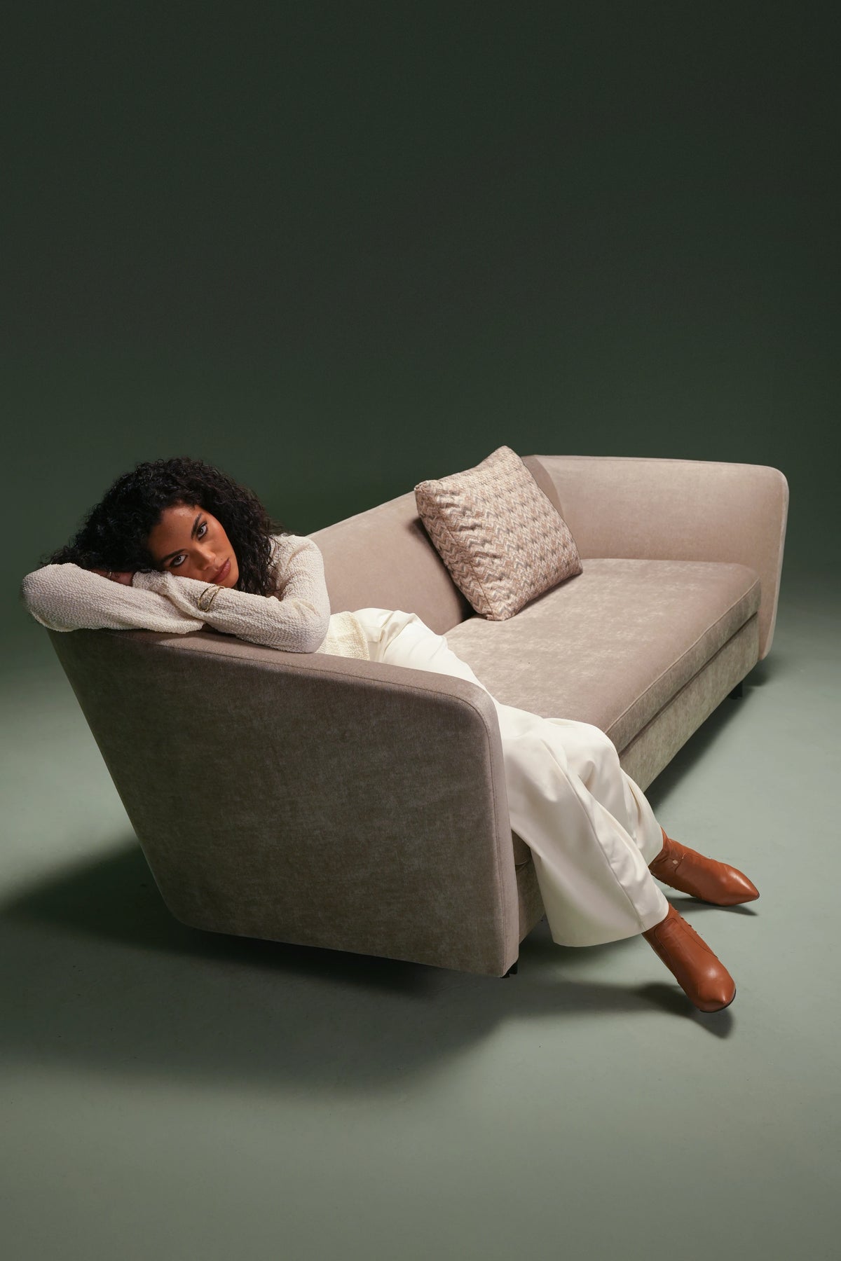 Woman resting on the beige Kirby sofa, showing its curved backrest transitioning from a higher side to a lower side.