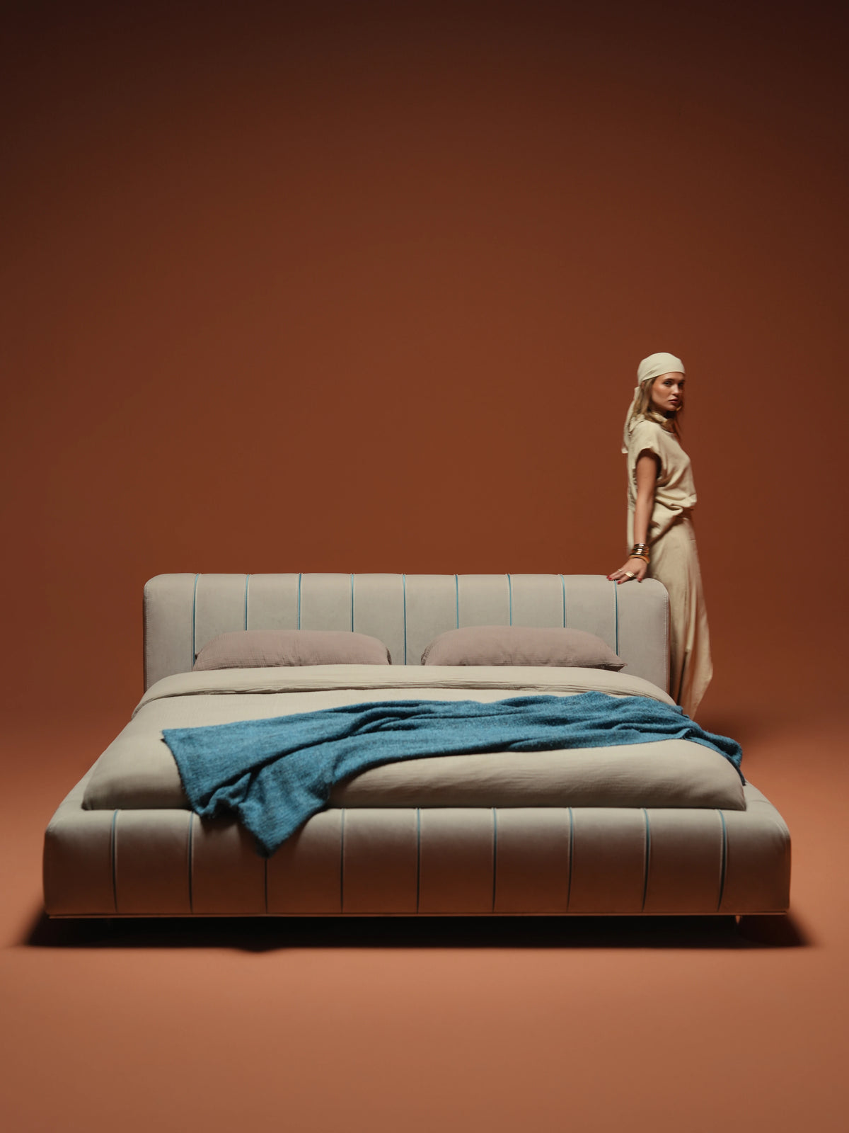 Woman standing behind the Lush upholstered bed with a recessed mattress, rounded beige frame and blue seam detailing.