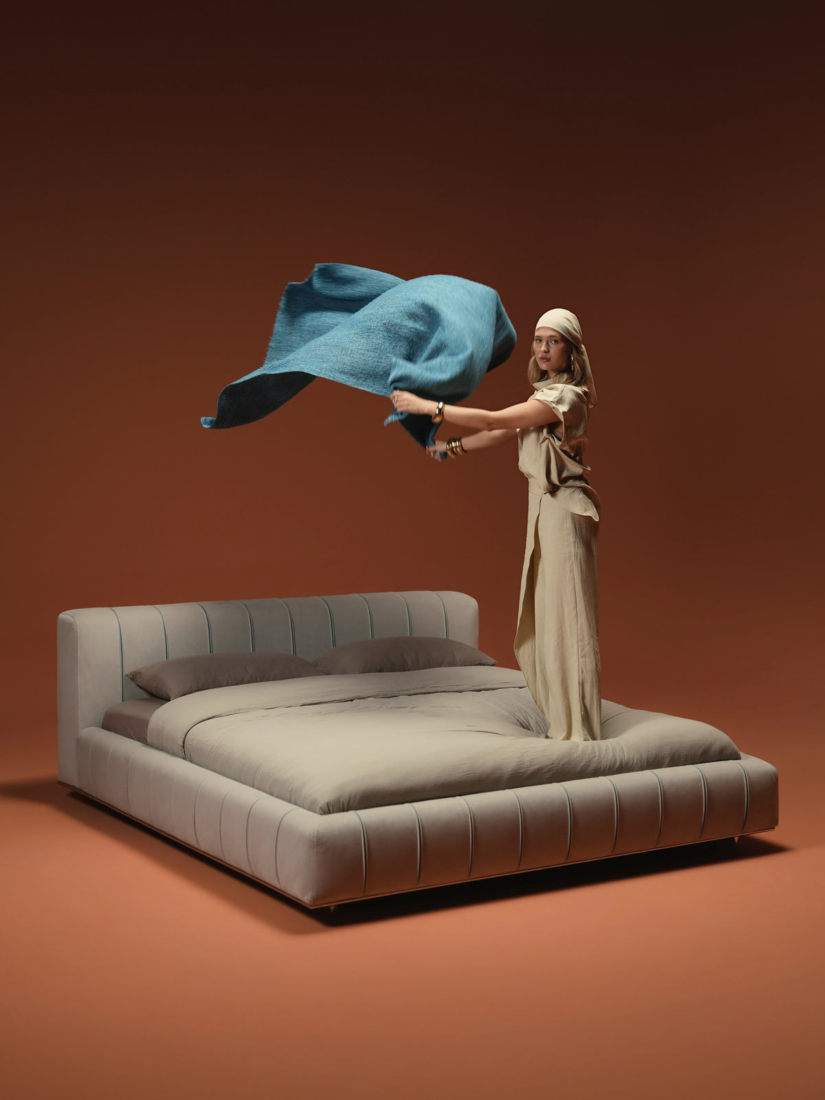 Woman standing on the Lush upholstered bed while lifting a blue throw, showing its recessed mattress and rounded beige frame.