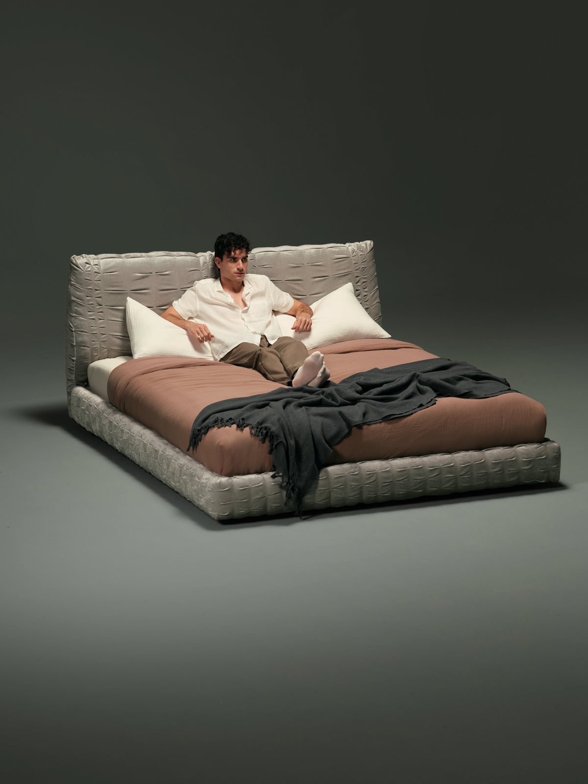 Man sitting on the Crusty upholstered bed with flexible pleated backrests, brown bedding and a dark grey throw.