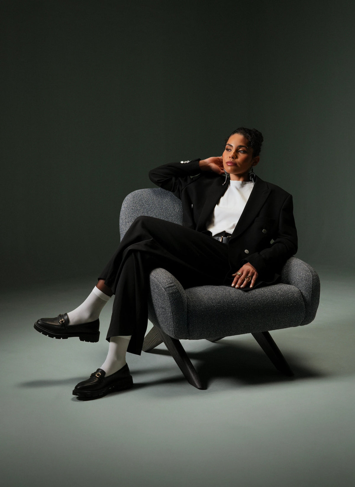 Woman seated in the grey Miska armchair, showing its curved upholstered arms, rounded backrest, and angled wood legs.