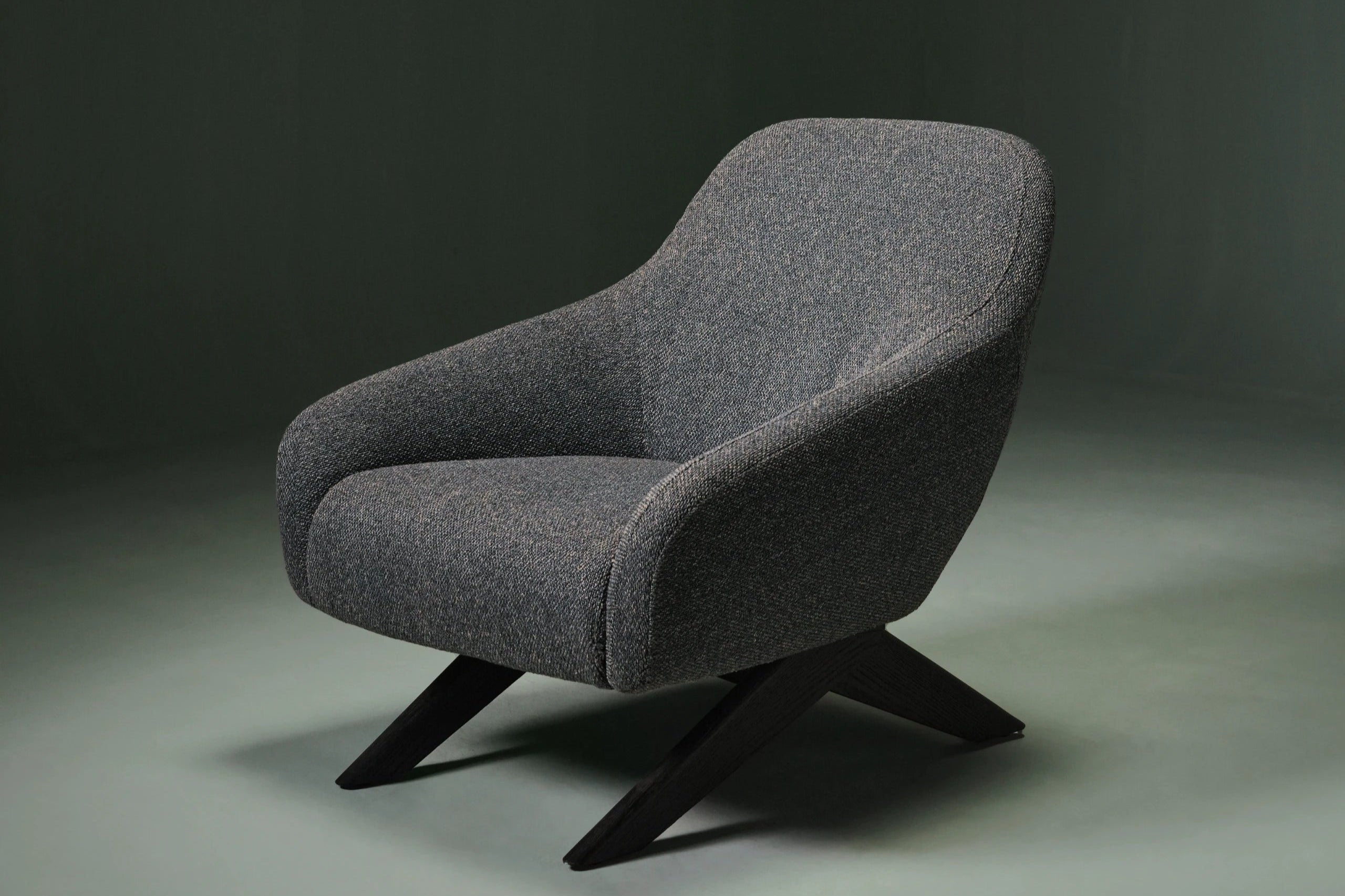 Miska armchair in textured grey upholstery, featuring a continuous curved shell and angled dark wood legs.