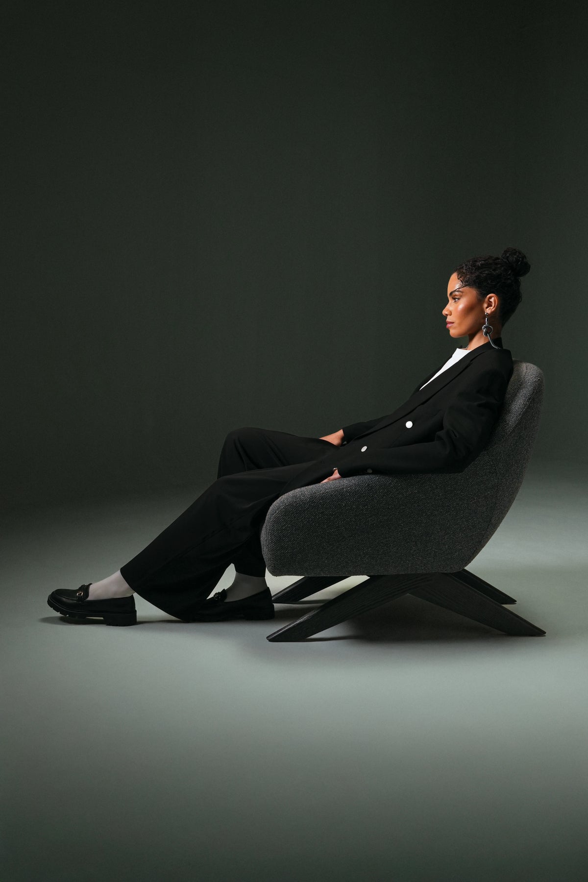 Side view of a woman seated in the grey Miska armchair, highlighting its continuous upholstered shell and angled wood legs.