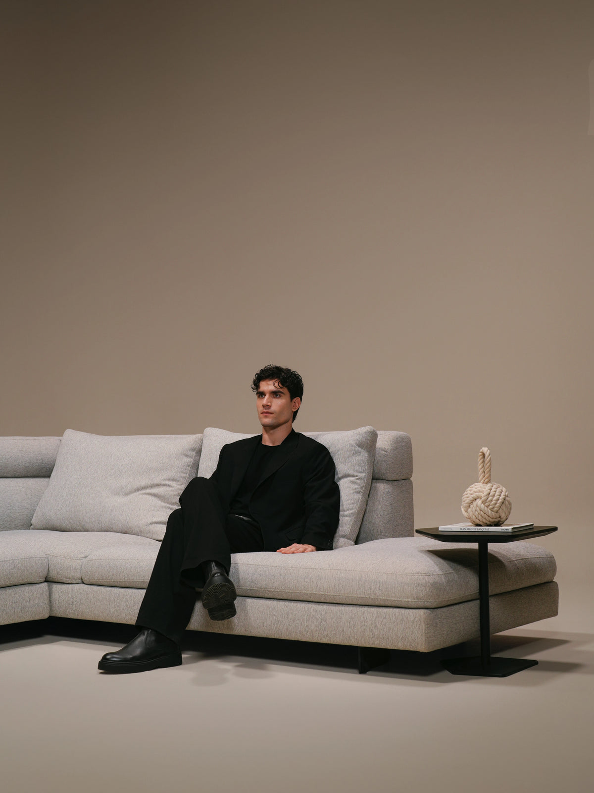 Man seated on the beige Monti sofa beside a slim side table, with deep seating and feather-filled back cushions.