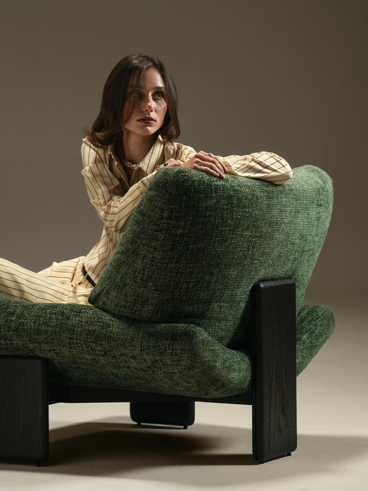 Woman seated in the green Muse lounge chair, shown from behind with its upholstered back and dark solid wood slab legs.