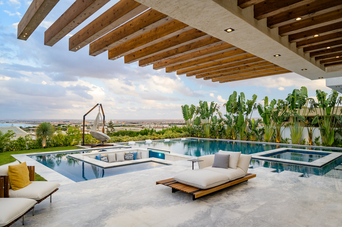 Nest outdoor chaise longue with an integrated accent table, with white cushions and teak platforms, arranged beside a swimming pool.