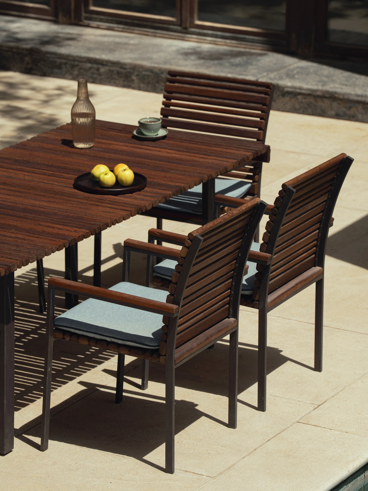 Close-up of Nest dining chairs with solid teak backrest and seat in alternating curvy slat widths, stainless steel frame with electrostatic coating, and cushioned seats upholstered for comfort, arranged around the matching table.