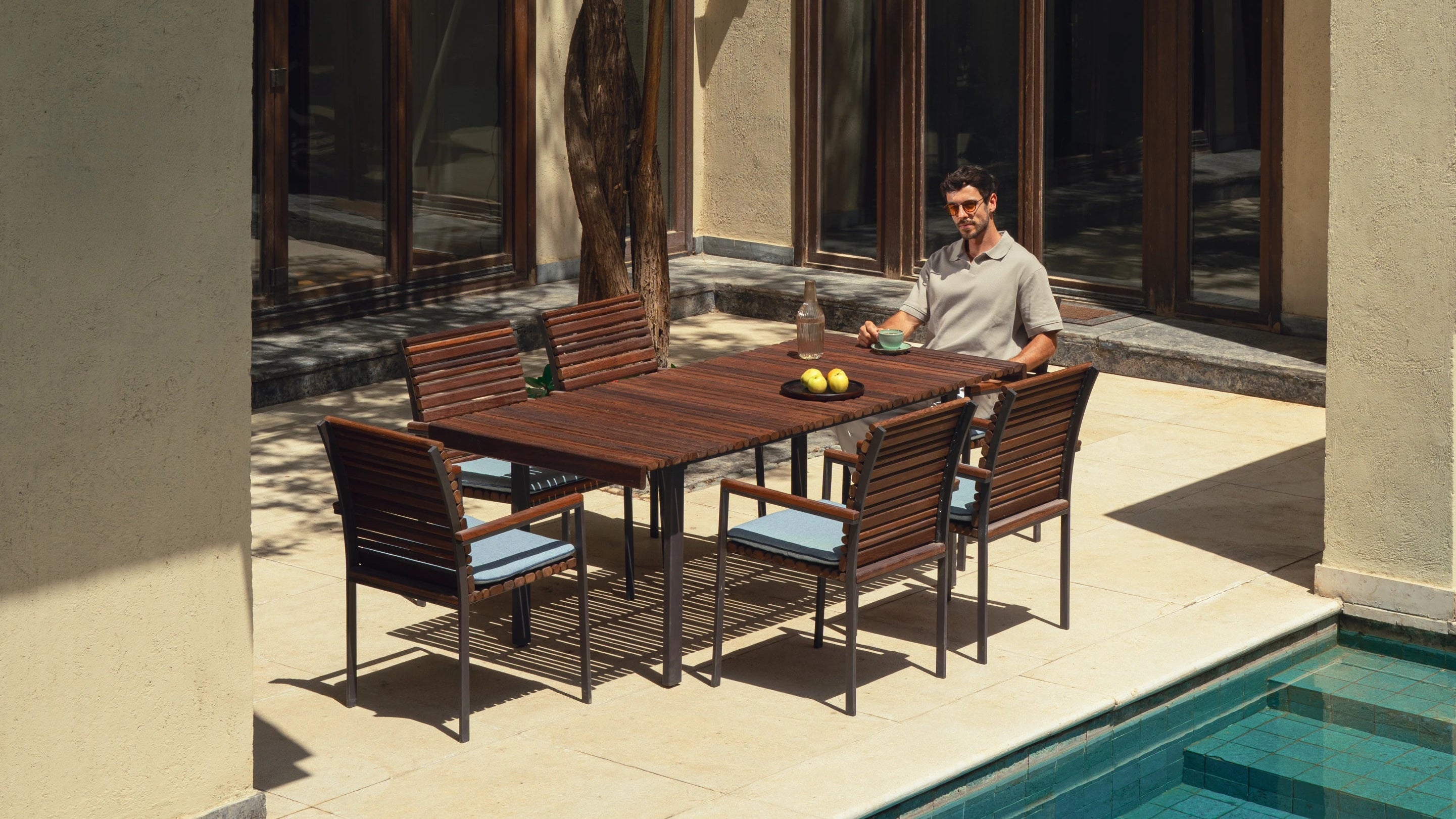 Nest dining table with solid teak top in alternating curvy slat widths, supported by a slim stainless steel frame with electrostatic-coated legs, paired with matching teak slat chairs and polyurethane cushions, styled poolside with model seated.
