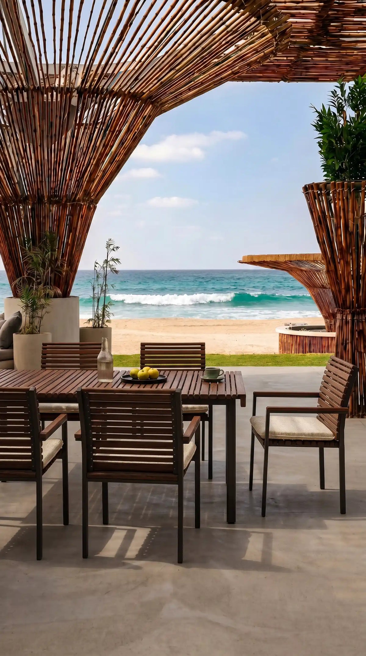 Nest outdoor dining set with a solid teak slatted table and matching cushioned chairs beneath a wooden pergola overlooking the sea.