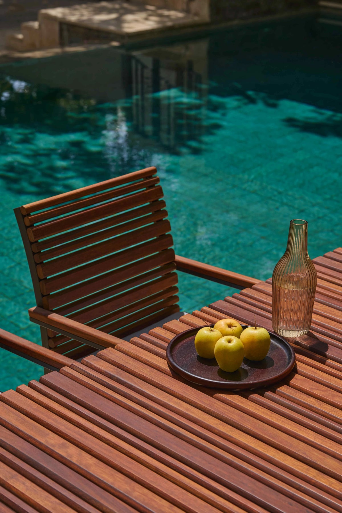 Close view of the Nest solid teak slatted dining table and matching chair beside a swimming pool, styled with green apples and a glass bottle.