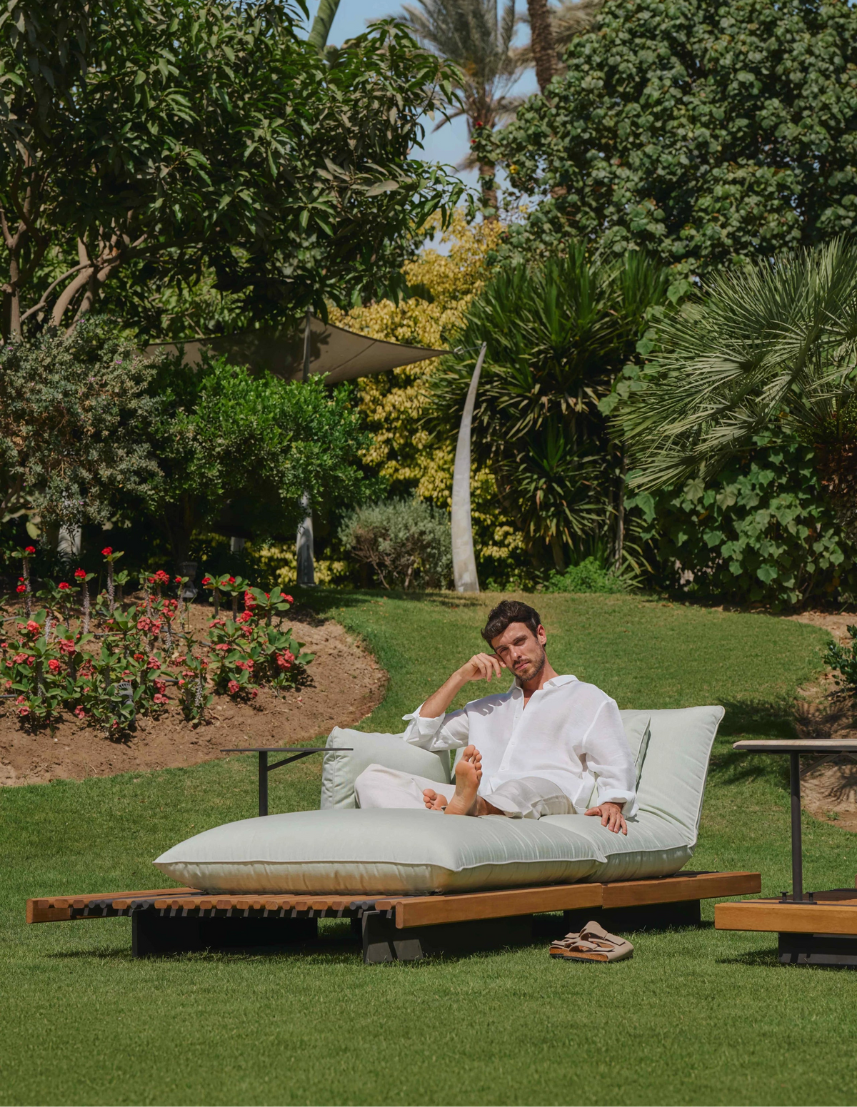Man sitting on the white Nest outdoor chaise longue beside an attached accent table in a garden.