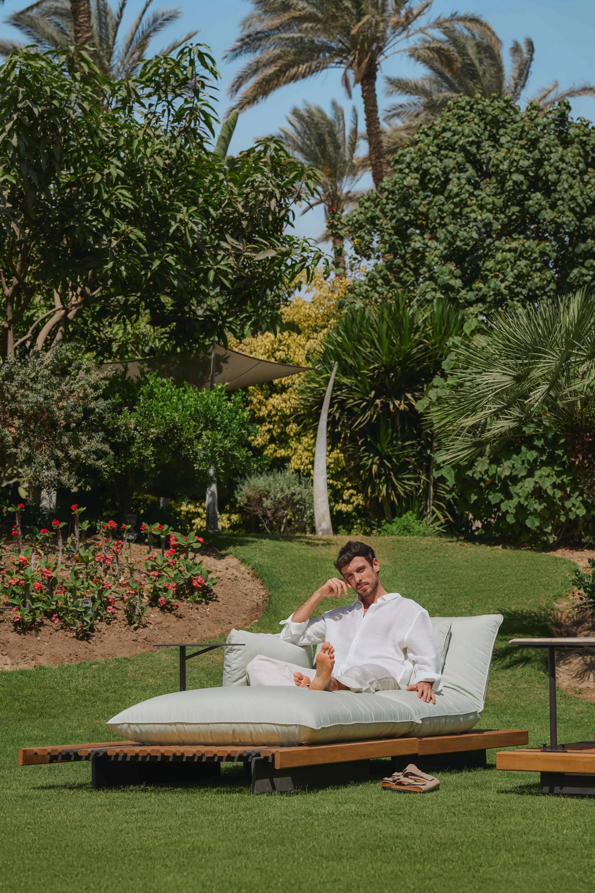 Man sitting on the white Nest outdoor chaise longue beside an attached accent table in a garden.
