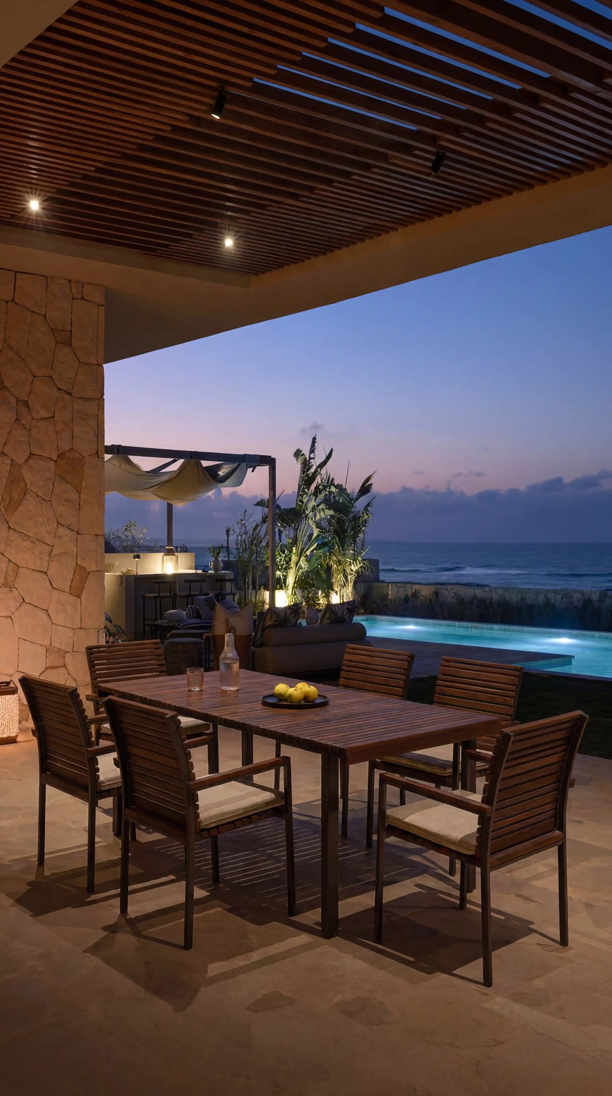 Nest outdoor dining set with a teak slatted table and six matching cushioned chairs on a covered terrace overlooking a pool and the sea at dusk.