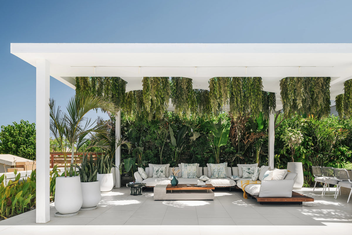 Nest outdoor sofa and table set with white cushions and teak platforms, arranged beneath a white garden pergola.
