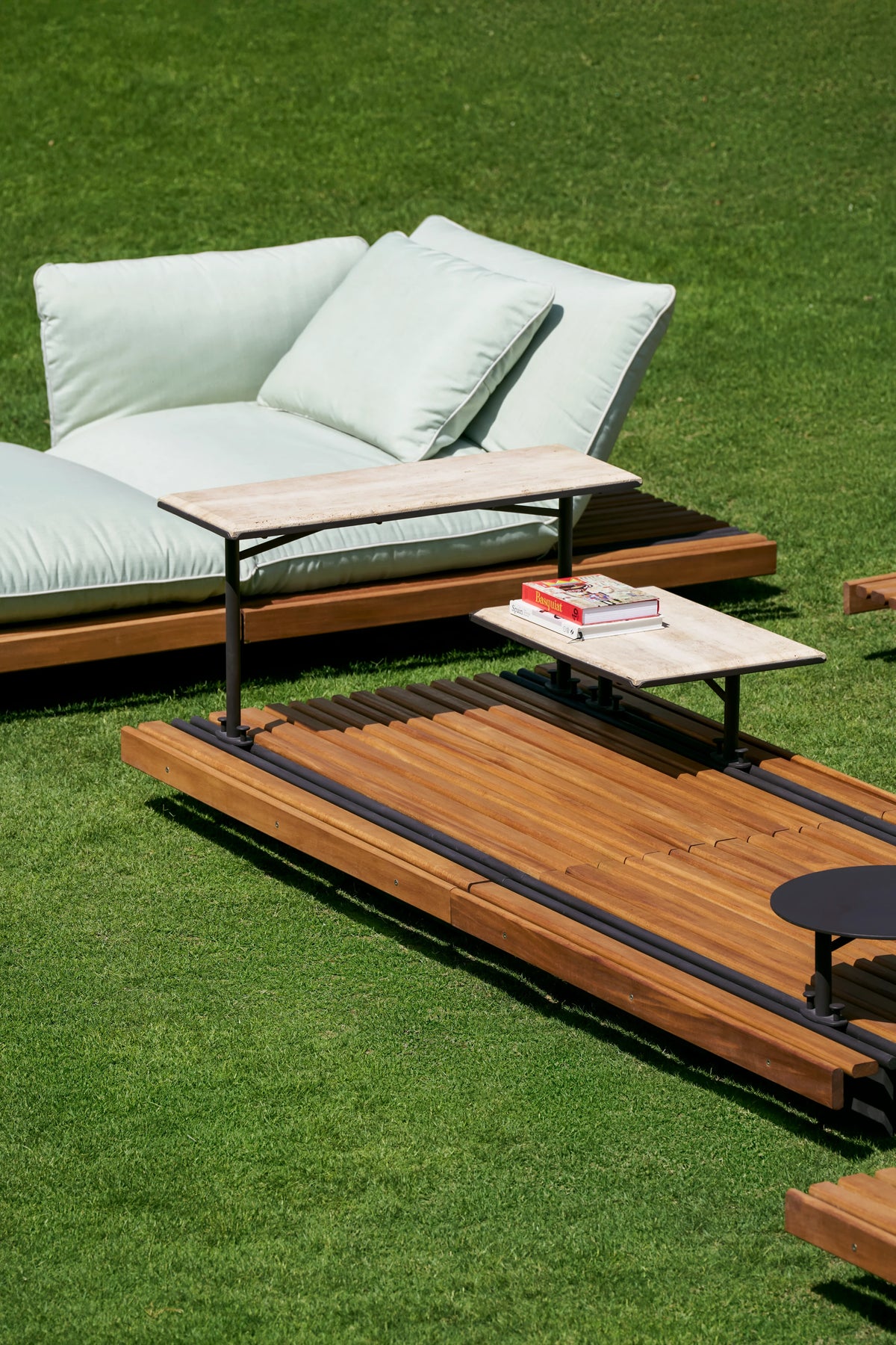 Nest outdoor table with mixed-width teak slats and raised accent tables, positioned beside the white Nest chaise longue on a lawn