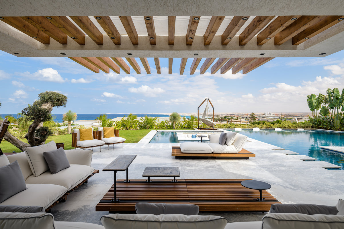 White Nest outdoor sofas arranged around the slatted teak Nest table beneath a poolside pergola.