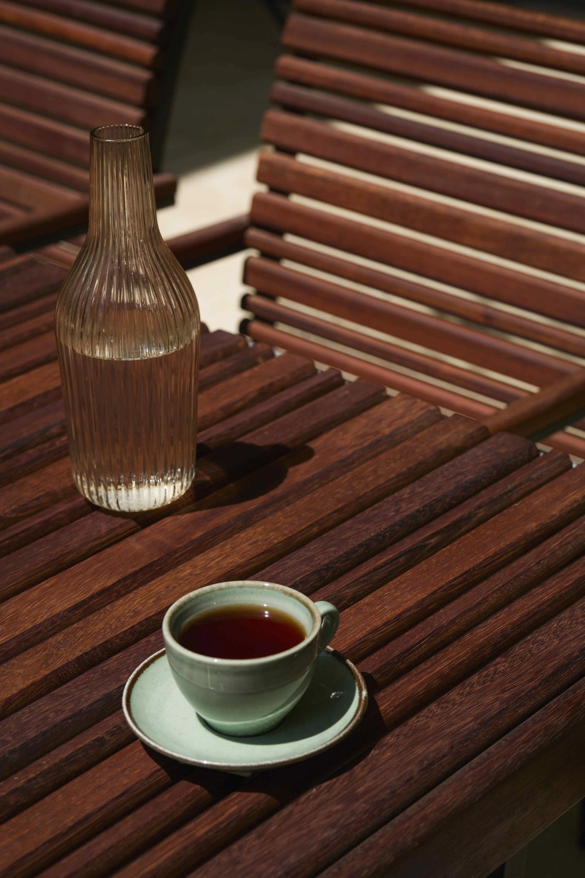 Close view of the Nest dining table’s solid teak top with alternating slat widths, styled with a glass bottle and teacup.