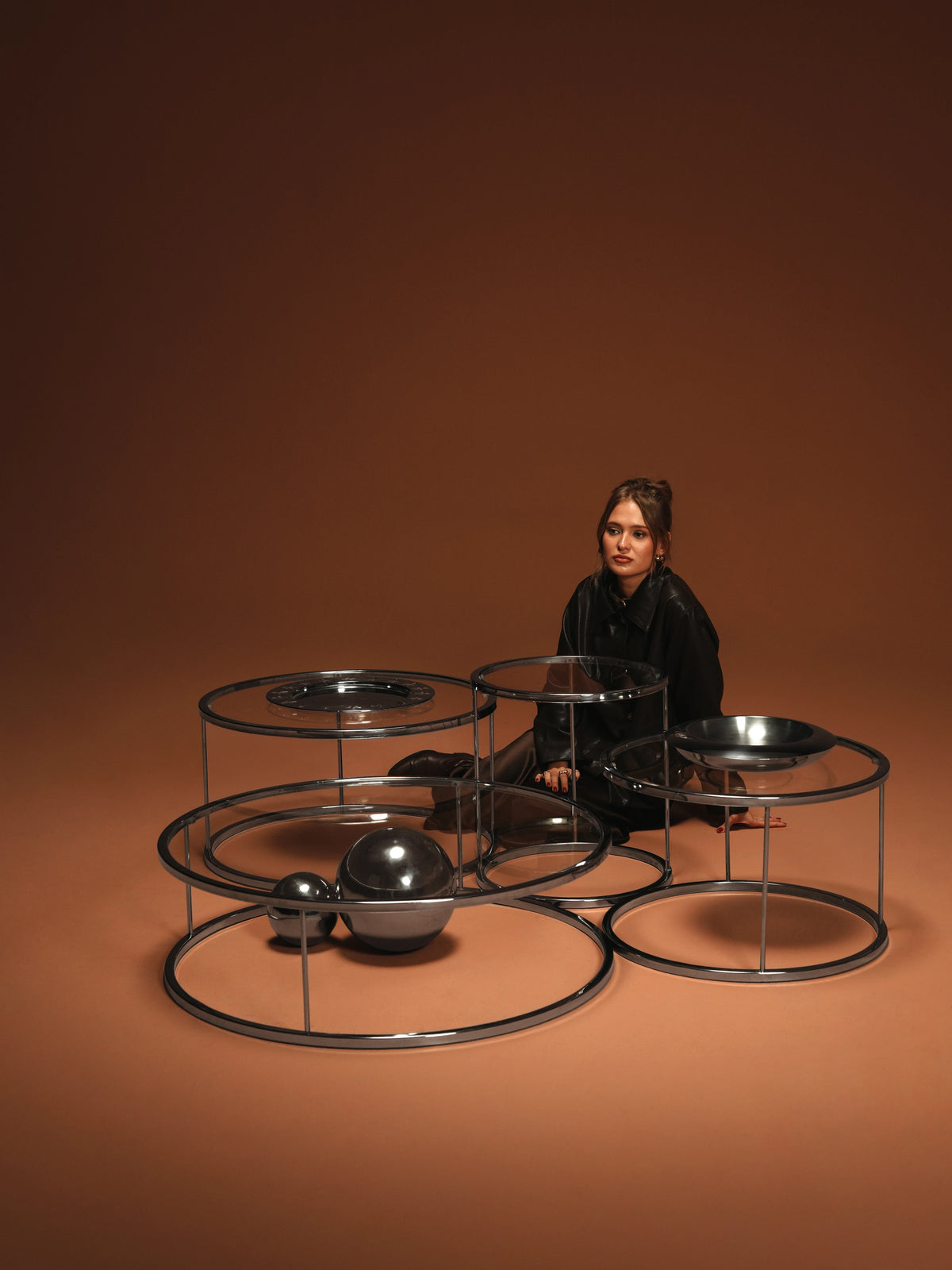 Woman seated among a grouped set of Orbit tables with round glass surfaces and polished stainless steel frames.