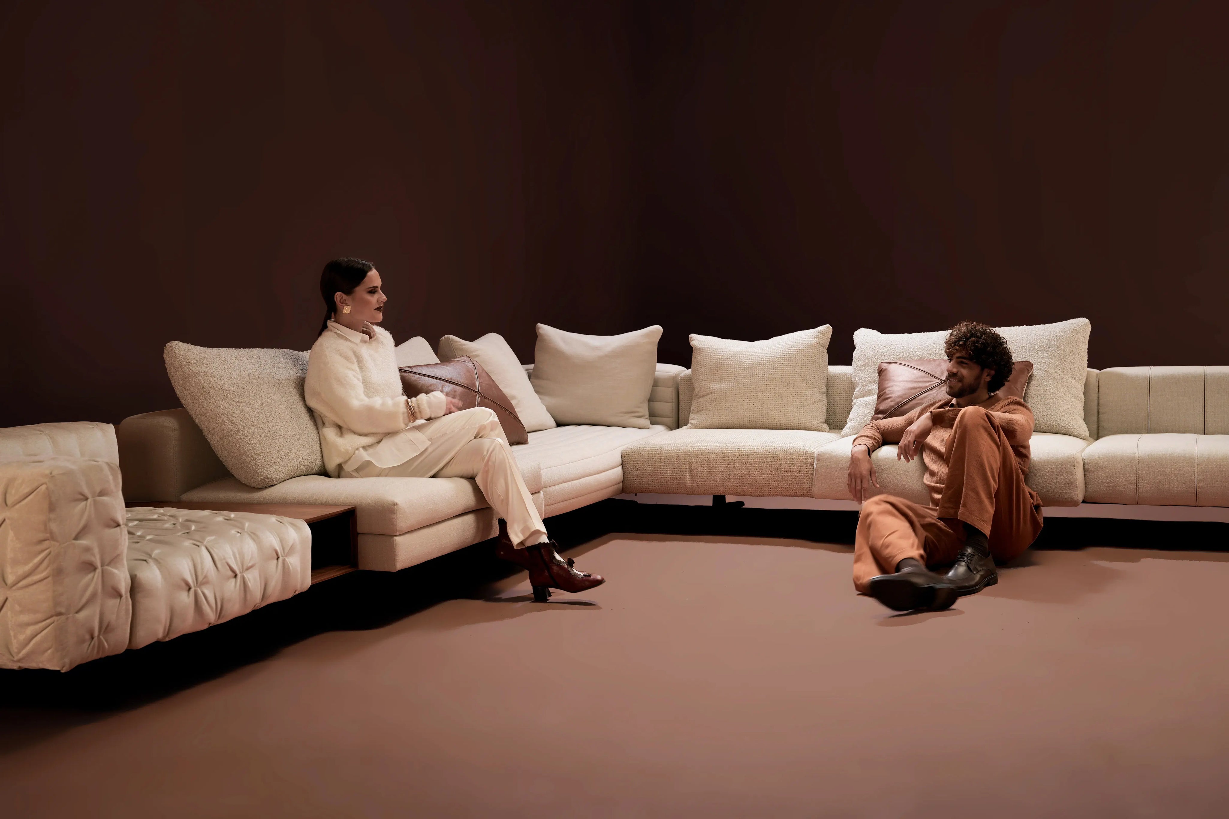 Two models seated around the Paella Sofa in a warm brown setting, showing its modular L-shaped design with plush cushions and mixed upholstery designs.