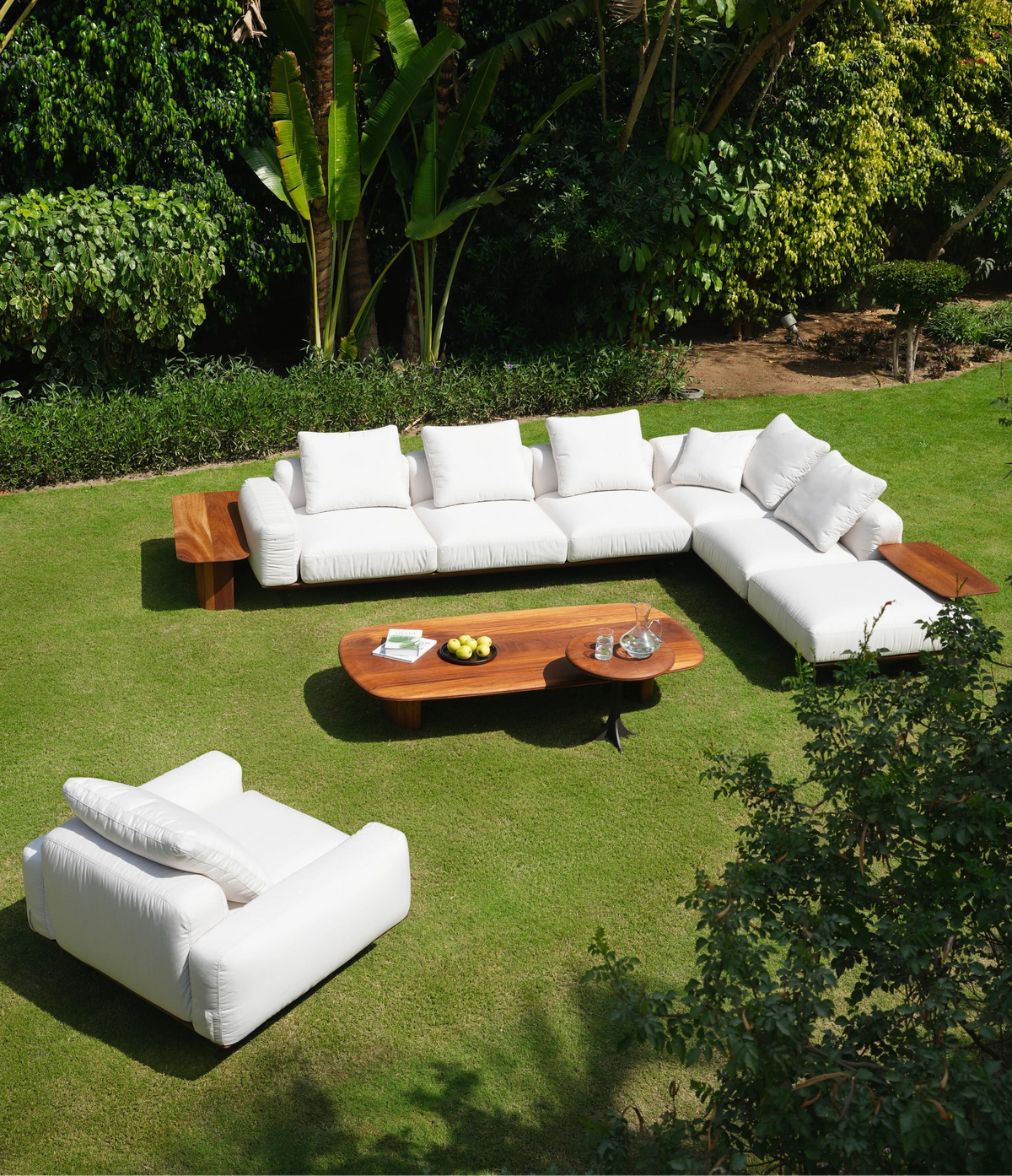White Puff sectional sofa and armchair arranged with large and small Glide tables and teak L Corner tables on a garden lawn.