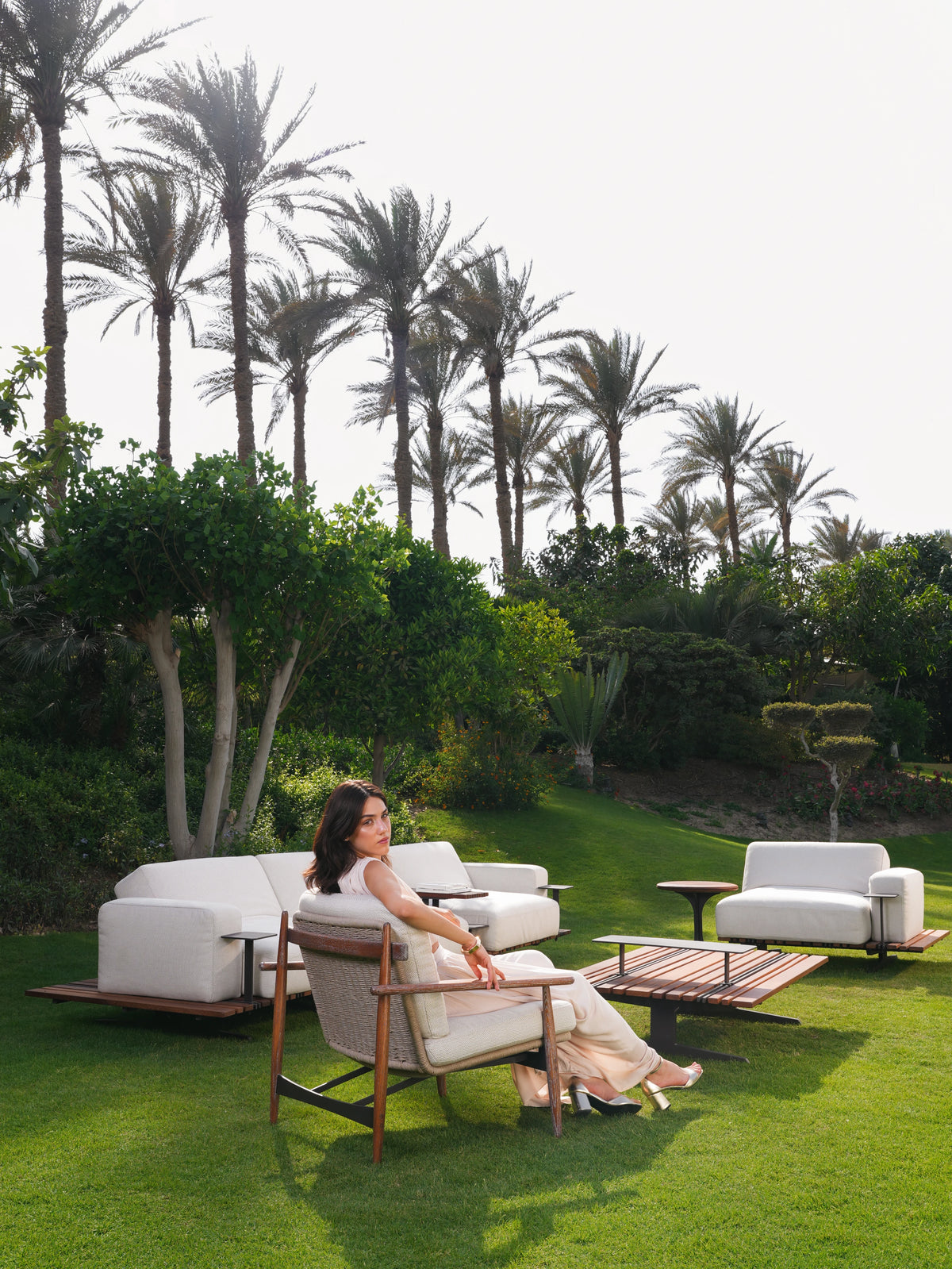 Woman seated in a Retro outdoor armchair with a wood frame and rope backrest, arranged with a Lodge sofa, armchair and slatted table in a garden.