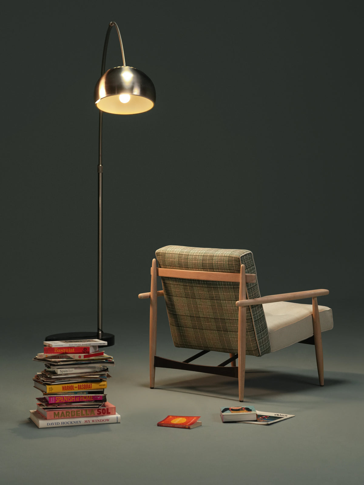 Rear view of the Retro Lounge armchair with a plaid upholstered backrest, solid wood frame, curved armrests, and a slim metal stretcher beside a floor lamp and books.