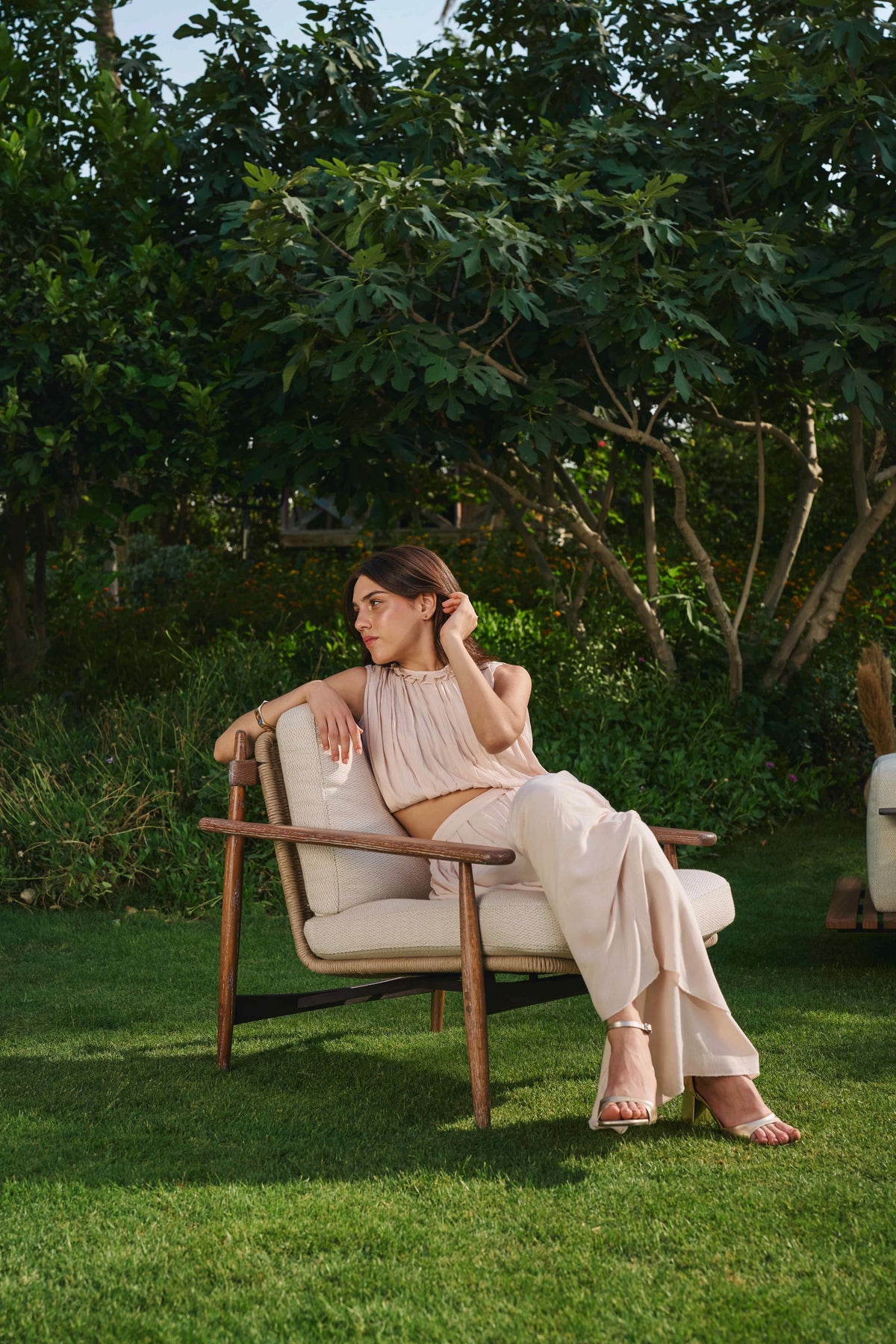 Woman relaxing in a beige Retro outdoor armchair with a solid wood frame, rounded armrests and woven rope backrest in a garden.