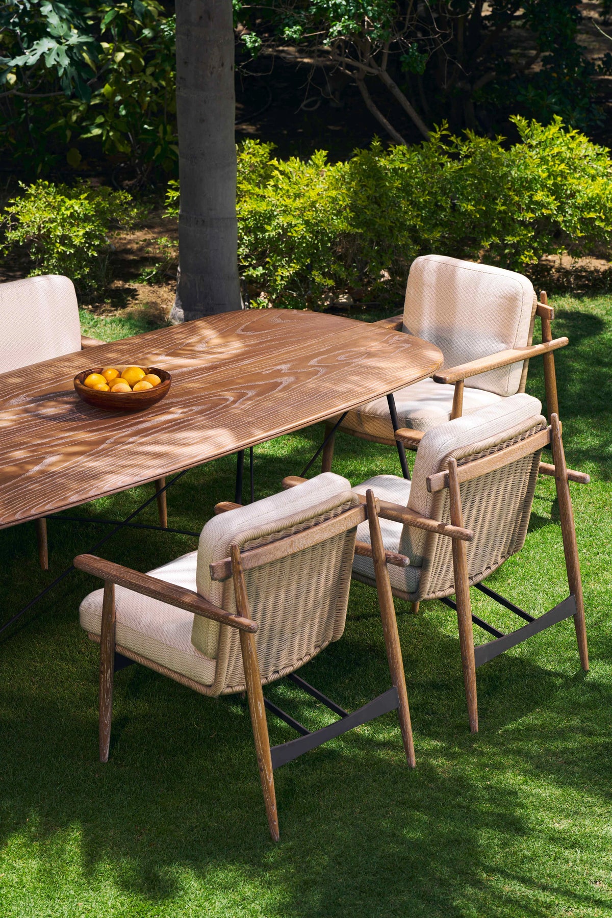 Retro outdoor dining set with a solid teak slatted table and cushioned wood-frame chairs with handwoven rope backrests, arranged on a garden lawn.