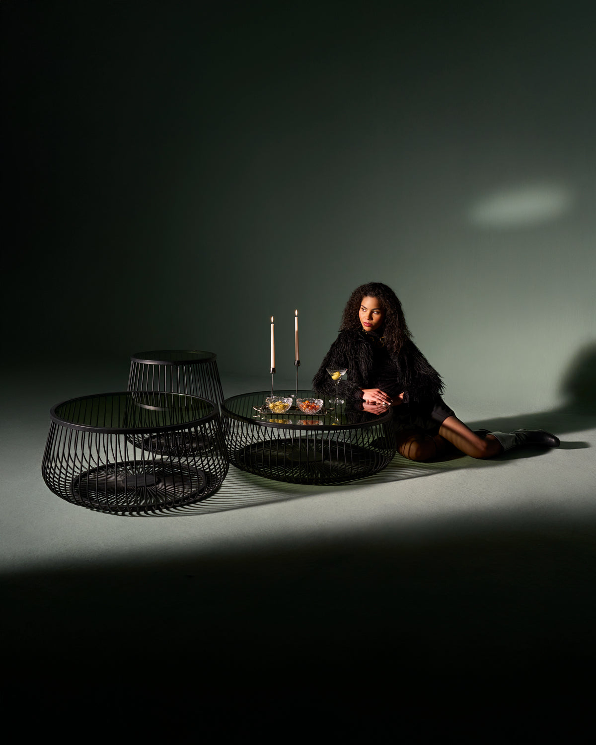 Model seated beside a Ring table with circular metal base and glass top, styled with candles and drinks.